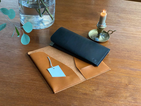 Two leather Portfolio Inserts, one saddle tanand one navy, on a wooden surface with a candle and plant in the background.
